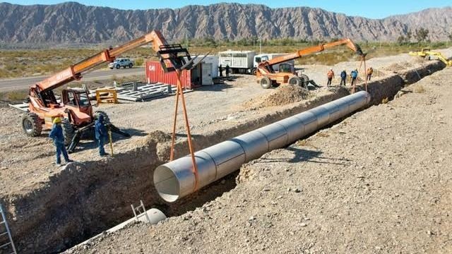 Trabajadores y maquinaria colocan una cañería en una zanja durante una obra de acueducto.