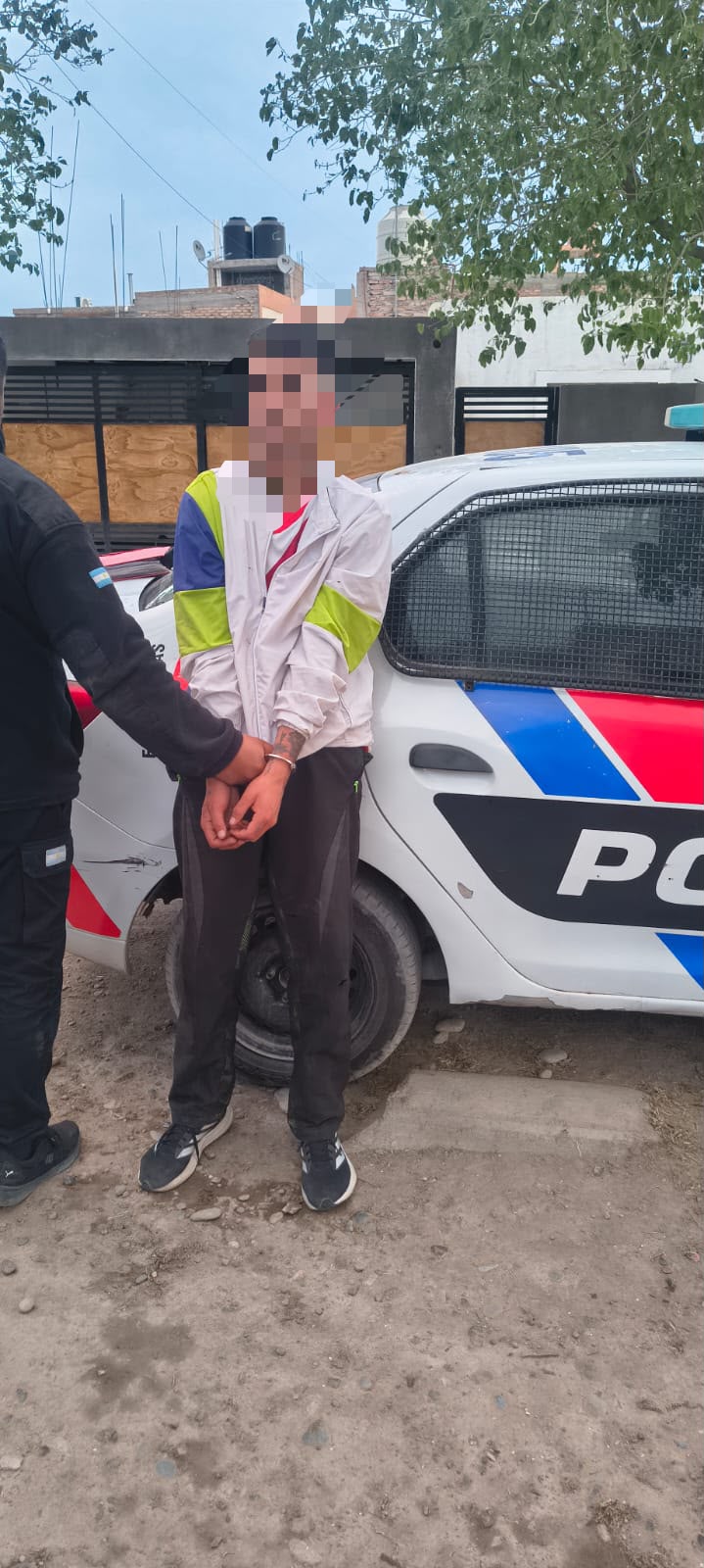 Hombre con rostro pixelado junto a un móvil policial, en la fotografía aportada sobre la aprehensión en el Lote Api II, Chimbas