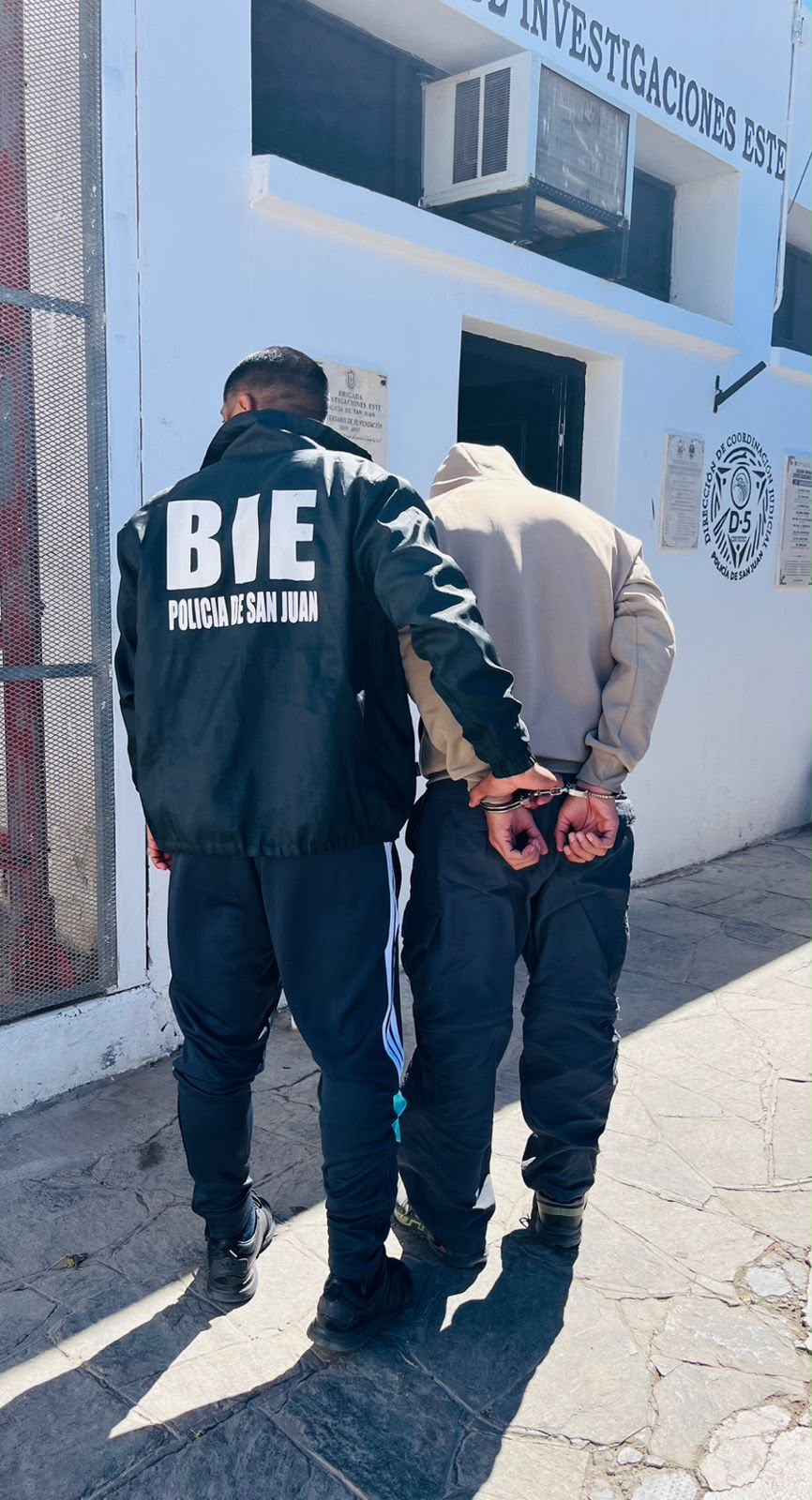Agente de la Brigada de Investigaciones Este junto a un hombre esposado, ambos de espaldas, frente a la dependencia policial
