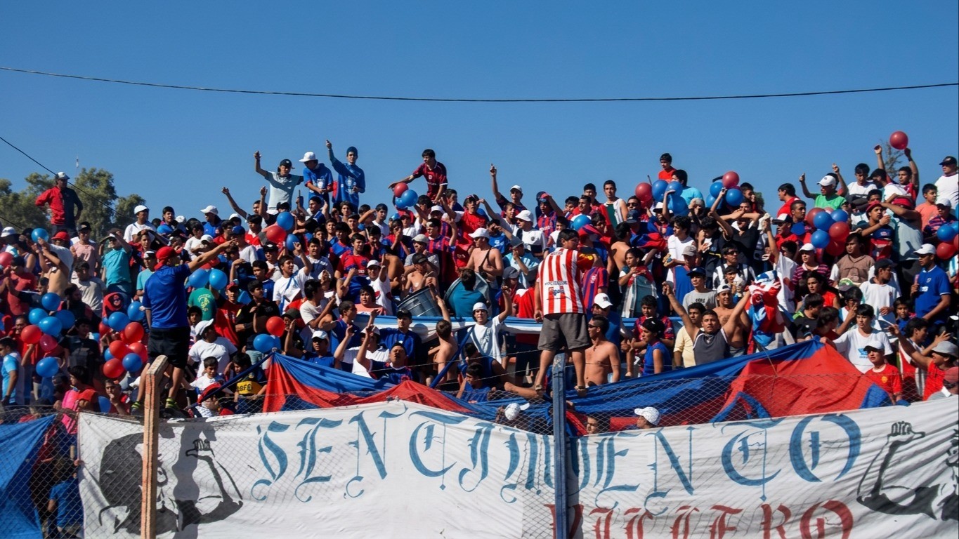 Hinchas de Villa Obrera en una tribuna con banderas rojas y azules