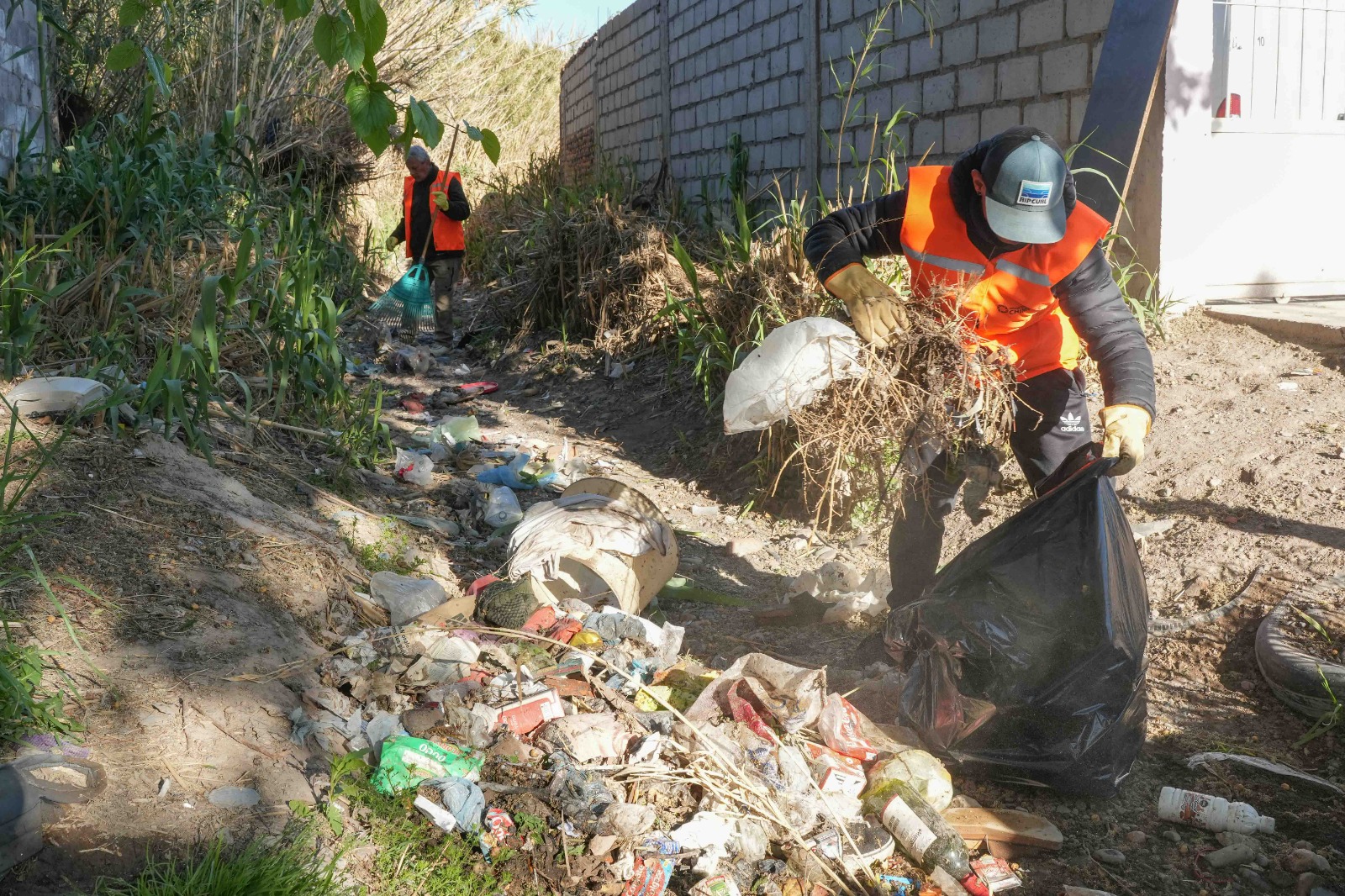 Un operario mete ramas en una bolsa mientras otro usa un rastrillo junto a una zanja con basura