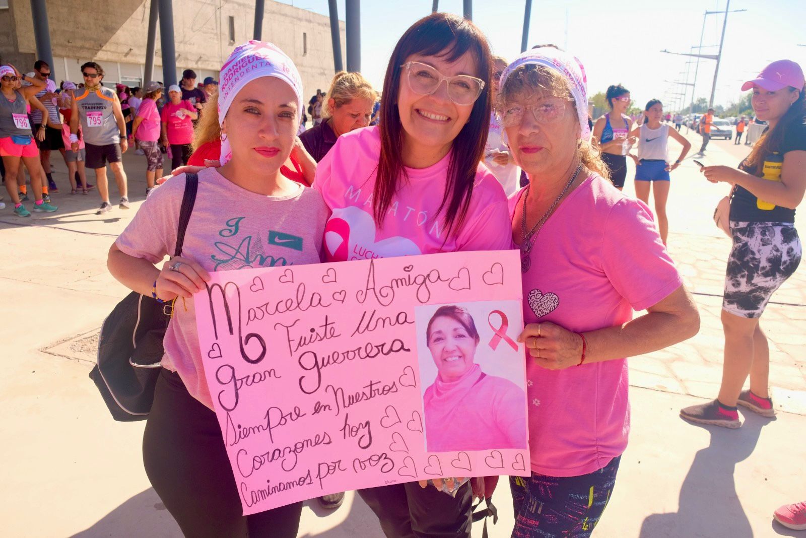 Tres mujeres sostienen un cartel rosa dedicado a Marcela durante la maratón