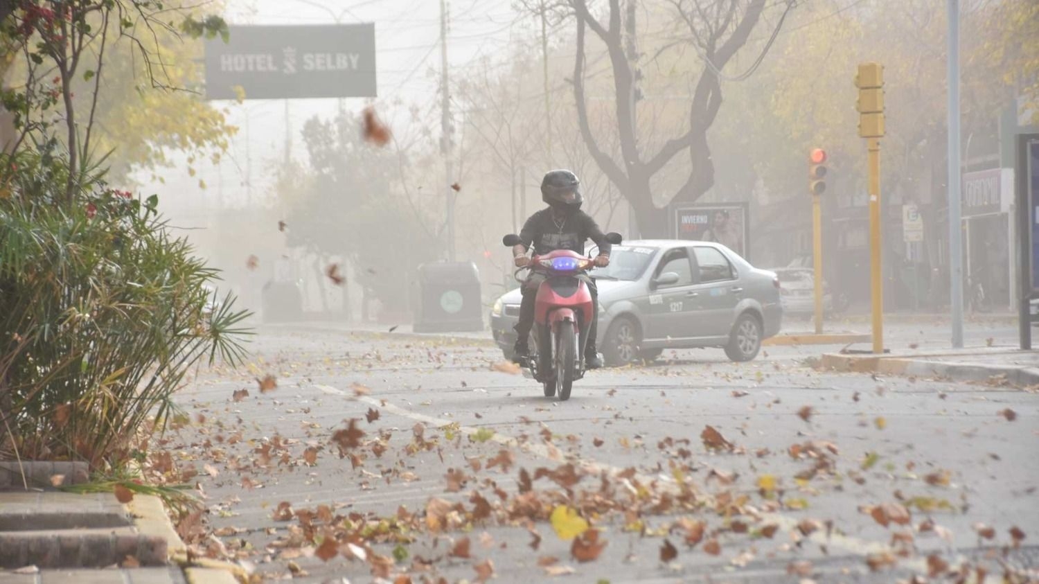 Una motociclista circula por una calle con polvo y hojas levantadas por el viento.