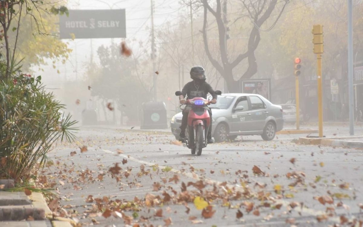 Una motociclista circula por una calle con polvo y hojas levantadas por el viento.